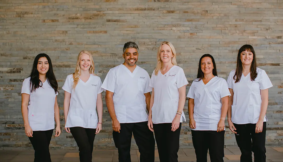 Equipo de dentistas y profesionales de Clínica Odontológica NOHE en Chicureo. Una imagen horizontal que muestra a un equipo médico de seis integrantes (cuatro mujeres y dos hombres) dispuestos en una fila. En el centro destacan David, un hombre de cabello oscuro con canas y barba corta, y a su lado Ranny, una mujer de cabello rubio largo. Junto a ellos se encuentra el resto del equipo. Todos visten camisas de uniforme blancas con el logotipo NOHE bordado y pantalones oscuros. El grupo transmite una actitud profesional y acogedora, posando con las manos a los costados o ligeramente al frente sobre un fondo de listones de piedra en tonos grises y tierra.